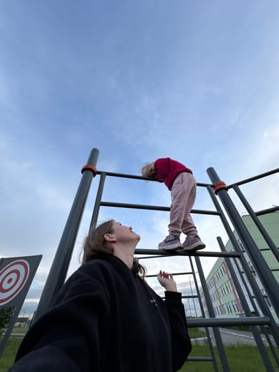 The image displays an outdoor scene featuring two individuals and a metal playground or exercise structure under a partly cloudy, light blue sky. In the lower-left foreground, an adult or teenager, with light brown hair and wearing a black hoodie, is looking upward, their right hand slightly raised with fingers loosely curled. Higher on the grey metal structure, a child wearing a dark pink long-sleeved top, light pink sweatpants, and light purple athletic shoes (Nike branding visible on the left shoe) is leaning forward over a horizontal bar. The metal structure incorporates orange-red rings on its vertical posts. To the left, a grey target board with red and white concentric circles is partially visible. In the mid-ground and background, a multi-story building with a light green facade, numerous windows, and white and orange horizontal stripe details is prominent. Green grass covers the ground in sections, and a paved area is visible. The scene is consistent with an urban recreational area, specifically located in the city of Горный, Russia.