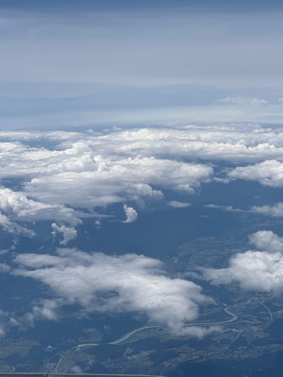 An aerial view captures a landscape from an airplane, dominated by a vast expanse of fluffy white clouds. Below the clouds, the earth is visible, showing a winding river with a turquoise hue. Along the riverbanks and surrounding areas, lush green terrain is interspersed with dark forests and scattered human settlements, indicating a rural or suburban environment. A road or highway network is visible, curving alongside the river and through the landscape, with some infrastructure and buildings visible. The sky above is a pale blue, fading into a lighter hue towards the horizon, with faint haze or distant clouds. There are no people or specific actions depicted in the image; it is a static, environmental scene. The crops suggest the image might be related to Glantschach, Austria, given the geographical features like the river and topography.