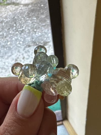A close-up shot shows a hand holding a small, clear beaded decoration. The decoration is clustered and appears to be shaped like a flower or a small animal, with several faceted beads reflecting light. The hand has a manicured nail with a French tip and a bright yellow accent on the cuticle area. In the background, a blurred outdoor scene with a light-colored, textured surface and hints of greenery is visible. The right side of the image shows a plain, light-colored wall and a dark vertical shadow. The scene is well-lit, suggesting it was taken during the day, possibly outdoors or near a window. The location of Naro-Fominsk, Russia, is not directly depicted or inferable from the visual elements in the image.