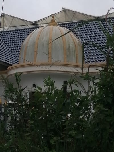A dome structure with golden accents sits atop a white building, partially obscured by lush green foliage. The dome is ribbed and appears to be made of a translucent material, possibly glass or plastic. Behind the dome, a dark, wavy tiled roof is visible, with a greenhouse-like structure with translucent panels extending upwards. The scene is outdoors, under an overcast sky. The overall impression is of a building with unique architectural features, possibly a home or a place of worship, situated in a verdant environment. The image does not depict any individuals or specific actions. The location in Lang Biang - Đà Lạt, Vietnam, suggests a potentially scenic and natural setting.