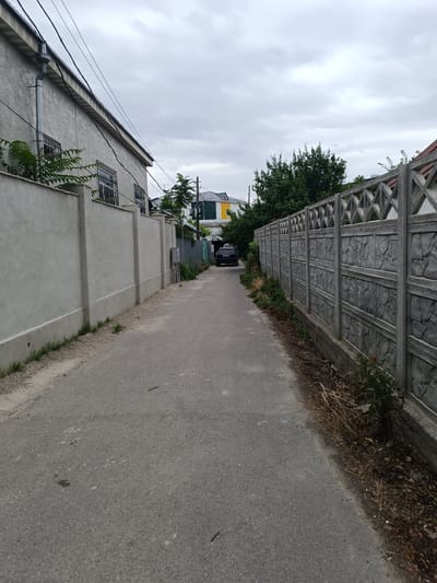 The image depicts a narrow asphalt alleyway in Taraz city administration, Kazakhstan, on an overcast day. A grey stucco wall runs along the left side of the alley, punctuated by barred windows and a downspout. A decorative concrete fence with a diamond pattern adorns the right side. In the distance, a dark-colored car is parked on the right side of the alley. The alley is lined with overgrown vegetation and dry grass on both sides. Further down the alley, glimpses of buildings with colorful facades can be seen. The sky is a dull grey, suggesting cloudy weather.