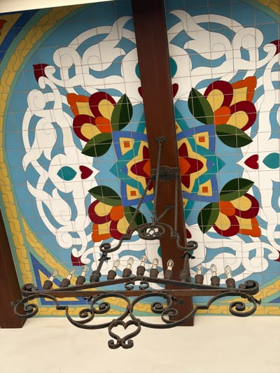 This image shows a close-up, low-angle shot of a wrought-iron chandelier hanging from a brown wooden beam. The chandelier has multiple light bulbs and ornate scrollwork. Behind the chandelier is a ceiling covered in colorful tiles, featuring a geometric pattern with floral and swirling motifs in blue, white, red, yellow, orange, and green. The tiles are arranged in a grid. Part of a white wall is visible at the bottom of the frame. The scene appears to be indoors, possibly in a building with an artistic or historical design, like the Huntington Library, Art Museum, and Botanical Gardens in San Marino, California, near Pasadena, United States of America, known for its decorative arts and architecture.