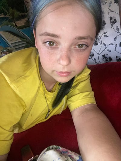 A young girl with blue-streaked hair is pictured in a close-up selfie. She is wearing a bright yellow t-shirt and a silver necklace. Her face is turned slightly towards the camera, and she has a neutral expression. Freckles are visible across her nose and cheeks. The background is blurred but appears to include a floral wallpaper and a red surface, possibly upholstery. To the left of the girl, a glimpse of an outdoor scene with palm trees and a blue structure, possibly a pool, can be seen. The location is not specified as Nova Praha, Ukraine.