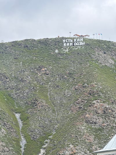 The image shows a mountainside with a sign in Russian reading "УСТЬ КАН КАН ООЗЫ". This corresponds to the city of Ust-Kan, Russia. The mountainside is covered in green vegetation and rocks. Above the sign is a structure that appears to be a viewing platform, with several flagpoles. The sky is overcast.