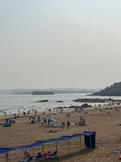 The image presents a wide, elevated view of a moderately populated sandy beach in Santander, Spain, under a hazy, light grey sky. The water is calm with gentle waves breaking near the shore, reflecting sunlight. In the mid-distance, the Faro de la Isla de Mouro lighthouse is distinctly visible on a rocky island within the bay, flanked by obscured, distant landmasses. The right side of the frame depicts a rocky coastline extending into the sea.
Numerous individuals are distributed across the beach and within the shallow waters. People are observed wading, swimming, standing, walking, and lying on towels or beach chairs on the sand. Several beach umbrellas, varying in color including blue, light blue, and green, are scattered across the midground of the beach, providing shade. In the foreground, along the bottom edge, are multiple interconnected blue fabric canopies supported by poles, shading rows of empty blue and white beach chairs. To the far right, underneath one of these canopies, a standalone blue fabric structure, resembling a changing booth, is present.
