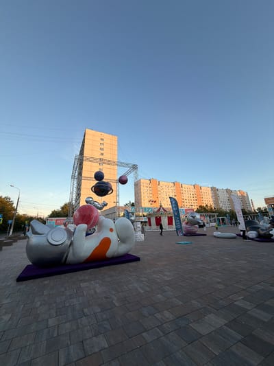 In Moskovskiy, Russia, an outdoor plaza is depicted with large inflatable sculptures. In the foreground, a silver and white inflatable figure resembling an astronaut is lying on its back on a purple mat. The figure has orange markings on its body and is holding a large red sphere. Behind the astronaut sculpture, a tall, light-colored apartment building stands prominently. Above this building, a metal structure, possibly part of a stage or exhibition setup, is visible. Several inflatable spheres resembling planets hang from this structure, some with rings. Further in the background, another long apartment building, this one with a more colorful facade, stretches across the scene. Various flags and banners are placed throughout the plaza. People are seen walking and standing in the background, indicating a public event or gathering. The ground is paved with large, rectangular tiles. The sky is clear and blue, suggesting it is daytime.