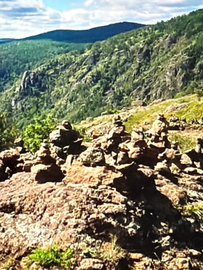 The image captures a mountainous landscape with a cloudy sky. In the foreground, there are several cairns, which are stacks of stones, placed on a rocky surface. The cairns vary in size and height. Behind the cairns, the terrain slopes upwards, covered with lush green trees and rocky outcrops. Further in the background, rolling hills covered in dense vegetation stretch towards the horizon, with a dark, undulating mountain range forming the backdrop. The overall impression is a natural, rugged environment. The provided location is Lopatino, Russia.