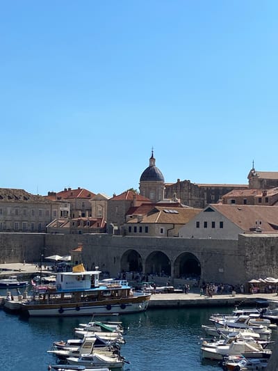 The image shows a harbor in Dubrovnik, Croatia, with the city walls and historic buildings in the background. The water is calm and blue, and several boats are docked. In the foreground, there are numerous small white boats. A larger white and blue passenger boat is docked to the left, with a yellow flag visible. Behind the boats, a stone promenade is bustling with people. The promenade is lined with arched openings, suggesting a passage or arcade. The buildings in the background are characterized by their stone construction and terracotta roofs, typical of Dubrovnik's architecture. A prominent domed building, likely a church or cathedral, is visible among the rooftops. The sky is clear and blue, indicating a sunny day. The scene captures a vibrant day at the harbor of this historic coastal city.