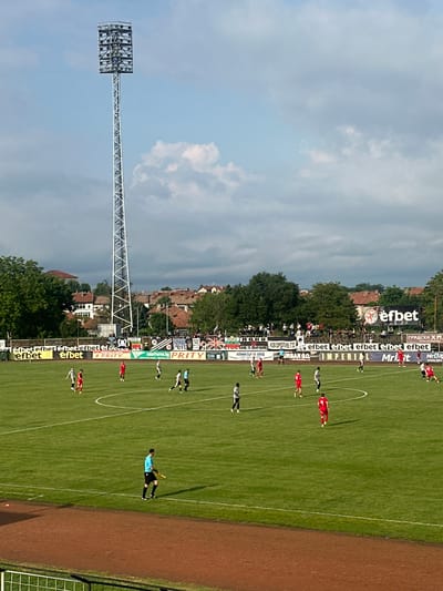 The image captures a soccer game in Gorna Oryahovitsa, Bulgaria. The view is from an elevated position, overlooking the field with players in red and white/black striped uniforms. A referee in a light blue shirt and black shorts is also visible. Advertising banners line the field, featuring brands like "efbet" and "PRITY," among others. The stadium has a low-rise brick building near the field perimeter. The sky is partly cloudy, and a tall floodlight structure is visible in the left part of the image. Spectators are present in the background.