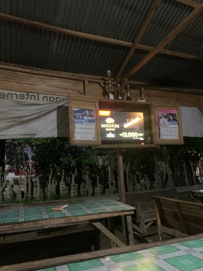 The image depicts an outdoor seating area, likely a warung or a local eatery, at night. In the foreground, there are several wooden tables with green and white tiled surfaces. An orange and white cigarette pack lies on one of the tables.

The background shows a structure with a corrugated metal roof supported by wooden beams. Mounted on the wooden structure are two framed calendars, one on the left and one on the right of a central television screen. The television displays an advertisement for "MAGNUM KRETEK" with a price of "Rp 15.000".

Behind the seating area, there is dense foliage and trees. A large white banner with text, partially obscured by the foliage, is visible on the left side of the structure. The overall impression is a casual, outdoor dining or gathering space in Kruengmanyang, Indonesia, illuminated by artificial light.