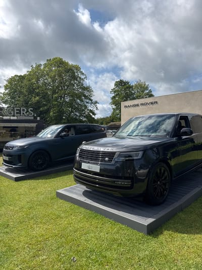 Two black Range Rover vehicles are displayed on raised platforms. The platform of the vehicle in the foreground is visible in the image. Both vehicles are facing the camera. Both vehicles have black alloy wheels and are parked on a grassy area. The vehicle in the foreground is a newer model. The vehicle in the background appears to be a slightly older model.  A person can be seen sitting in the driver's seat of the background vehicle, looking out the front window. The background vehicle has a "Range Rover" badge on the tailgate. The vehicle in the foreground is labeled "Range Rover" in silver letters on the front grille. The vehicle in the foreground has a "Range Rover" badge on the front grille,  a black license plate, and a black plastic plate below the license plate that says "Defender". Both vehicles are parked in front of a white building. The building has "Range Rover" in black lettering on the exterior wall. There is a food truck in the background. The food truck has the word "Burgers" on the side. The image was taken on a partly cloudy day with a blue sky. The photo was taken in the city of Bramham, United Kingdom.