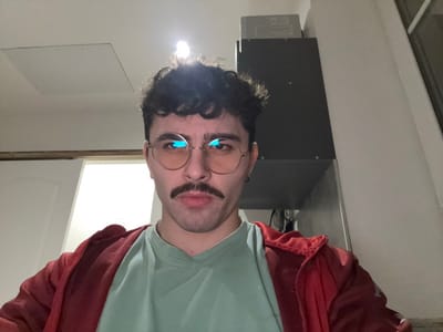 A person with curly dark hair, glasses, and a mustache is facing the camera. They are wearing a light green t-shirt and a red jacket. Behind them is a gray cabinet or appliance on the left, and a skylight in the ceiling towards the right. A square access panel is also visible in the ceiling. The image is oriented upside down.