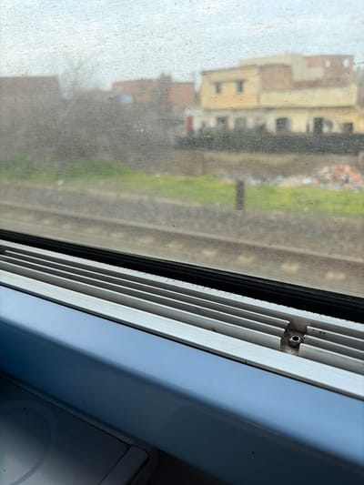 The photo captures a view through a train window. Outside, blurred buildings and a strip of green vegetation are visible. The window itself is quite dirty. The window frame, a white, slatted design, dominates the right side of the frame. It has a gray, slightly corroded bracket holding the slats together. The window frame is a very bright white.