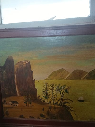 A framed landscape painting is centrally depicted, positioned beneath a white-framed window, in the city of Dogsenang Gorphoog, Bhutan. The painting, an oil on canvas, features a sky transitioning from light blue-green at the top to a muted orange-yellow horizon line. Below the horizon, a large body of olive-green water occupies the central and right portions of the image, bordered by multiple rounded, green-brown hills or mountains in the distance. Two small, dark boat-like forms are visible on the water in the mid-distance, and a larger sailboat with a dark hull and white sails is positioned in the lower-right quadrant of the water.
The foreground comprises a brown-toned landmass. On the left, two prominent, dark brown rock formations or cliffs rise; the leftmost is slender and conical, while the central one is wider with a relatively flat top and irregular, jagged sides. A small, white house with a dark roof and a single dark window is situated in the lower-left, partially behind the rock formations, with a faint white fence in front. Two small, dark, indistinct human figures are present on the land; one has a small reddish detail and is near the house, while another dark figure is situated further right. Scattered trees, including slender dark-branched forms with sparse green foliage and a denser cluster on a promontory extending into the water, are visible. Several dark, irregular rocks are on the ground in the lower-center. A faint dark signature or inscription is located in the painting's bottom right corner.
Above the painting, a bright, diffused light source is visible through a white-framed window, which exhibits minor weathering. A small, light-colored piece of debris or paper is affixed to the upper right portion of the window frame.