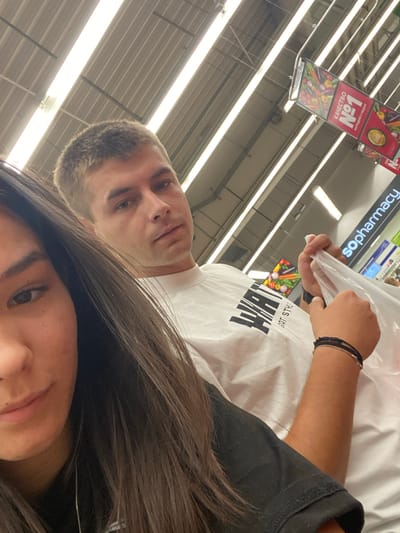 A close-up selfie shows a young Caucasian couple inside a brightly lit supermarket. The woman is on the left, with her dark hair partially covering her face, and the man is on the right, wearing a white t-shirt. He is holding a clear plastic bag in his right hand and has a beaded bracelet on his wrist. In the background, a sign with vegetables and the text "КАЧЕСТВО №1" (Quality №1) is visible, along with "sopharmacy" branding, indicating the location is likely a Kaufland supermarket in Sevlievo, Bulgaria. The ceiling features fluorescent lighting and metal structures, typical of a commercial interior.