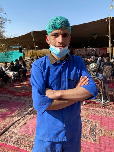 This image depicts an outdoor gathering in Sard-E 'olya, Afghanistan, under clear blue skies with bright sunlight. The foreground is dominated by a young adult male, positioned slightly left of center, looking directly at the camera with a neutral expression. He wears a teal-green disposable hairnet and a light blue surgical mask pulled down below his chin. His attire consists of a blue work shirt with a dark brown collar and a left chest pocket, and matching blue trousers. His arms are crossed over his chest.

The ground is covered with multiple red and black patterned rugs or carpets. In the mid-ground and background, numerous individuals, primarily males, are seated on chairs or on the ground, attending an event. They are dressed in various casual and traditional garments. A large, makeshift canopy constructed from camouflage netting or similar fabric, supported by poles, provides shade over the gathering. A lighter-colored tarp is visible further left. Bare tree branches are present on the far left. A banner or display board with red and white text and graphics is visible under the main canopy.