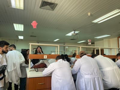 Inside a bright laboratory classroom, students wearing white lab coats are gathered around lab benches, seemingly engaged in an experiment. A woman is positioned behind a lab bench, smiling and gesturing with her hand. The room has fluorescent lighting, visible ceiling fixtures, and a projector. Decorative paper cutouts are hung from the ceiling. Lab equipment and personal belongings are visible on the benches.