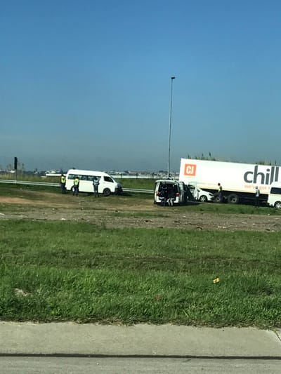 A white truck with "chill" printed on the side and a smaller white truck are parked off the side of the road with people nearby. The surrounding area is green grass and dirt, with a concrete sidewalk visible on the far right. A tall pole stands in the distance under a blue sky. Buildings are faintly visible in the background. The photo is taken at an angle, giving a tilted perspective.