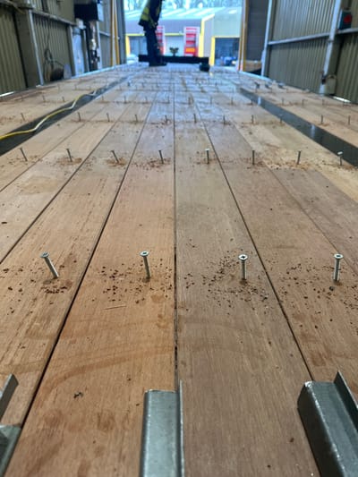 This image, taken in an industrial workshop in Lancaster, United Kingdom, presents a low-angle perspective of a large wooden deck, likely a vehicle or trailer bed, undergoing construction or repair. Numerous silver, self-tapping screws are partially driven into the light-brown wooden planks, with wood fiber debris scattered around their insertion points. In the immediate foreground, two parallel galvanized metal C-channel sections are visible. A yellow electrical cable runs along the left side of the wooden surface. In the distant background, a person wearing a high-visibility yellow jacket and dark trousers stands facing away from the camera, near the far end of the wooden structure. Behind the person, a large bay door is open, revealing an exterior environment with green foliage and the walls of other industrial buildings, some featuring red-orange bay doors, under natural ambient light. The interior walls of the workshop are clad with corrugated metal panels.
