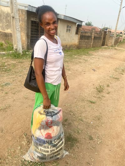 A woman stands on an unpaved dirt road in Odo Ona, Nigeria, facing slightly left of the camera and smiling broadly. She has dark skin, dark hair tied up, and wears a white short-sleeved t-shirt with a blue graphic, green sweatpants, and a black shoulder bag. A gold-colored bracelet is on her right wrist. She holds a large, translucent woven plastic sack with her right hand, visible contents include various items. Text on the sack reads "INDIANA CHEF," "100% INDIAN PARBOILED LONG GRAIN 5% BRISURE," "QUALITÉ SUPÉRIEURE," "POIDS: 50 KGS," and "FOOD FOR HUMAN CONSUMPTION." Behind her to the left is a single-story building with a light-colored wall, a barred window, and a corrugated metal roof. A concrete utility pole is visible near this building. Further right, a concrete block wall fence with a metal gate and another building with a corrugated metal roof and an antenna are present. The ground is dry, light brown, with sparse patches of dry grass. The sky is a light, hazy grey-white.