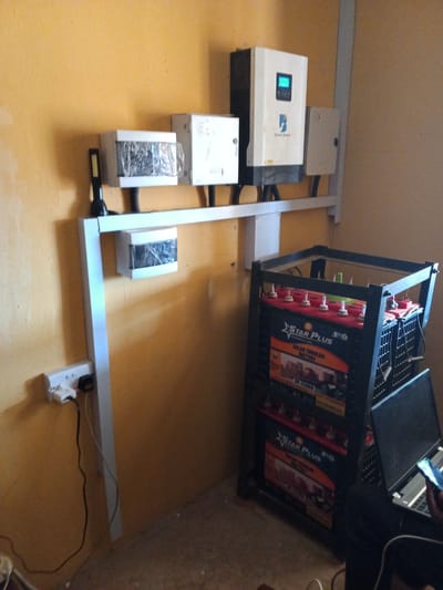 In an indoor setting with orange-painted walls and a brown floor, a solar power system is set up. The system includes multiple components mounted on a metal rack system attached to the wall. At the top left, a small flashlight is attached to the rack. To its right are two white boxes, one with plastic wrapping. Further to the right, a larger white inverter with a blue screen displaying text and a logo is mounted. Next to the inverter is a small, light-colored electrical box. Below these components, the metal rack extends horizontally, supporting another white box with plastic wrapping.
On the right side of the image, a black metal rack holds several large batteries. The batteries are black with red tops and feature the "STAR PLUS" branding and "SOLAR TUBULAR BATTERY" text. Some batteries are stacked on top of each other. To the right of the batteries, a black laptop is open on a surface, and a hand holding a smartphone is visible in the foreground.
On the left wall, near the floor, a white electrical outlet with a black power cord plugged into it is visible. The overall scene suggests a functional solar power setup in what appears to be a residential or small commercial space. The location of Kaduna South, Nigeria, is inferred from the provided context.