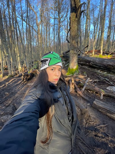 A young woman poses for a selfie outdoors in a wooded area. She is wearing a green, white, and black patterned beanie, a black puffer jacket, and a dark-colored outer jacket. Her hair is long and dark. The background consists of thin trees with bare branches, silhouetted against a bright sky. The ground is covered in dirt, roots, and fallen logs. The location is Unknown, Argentina.