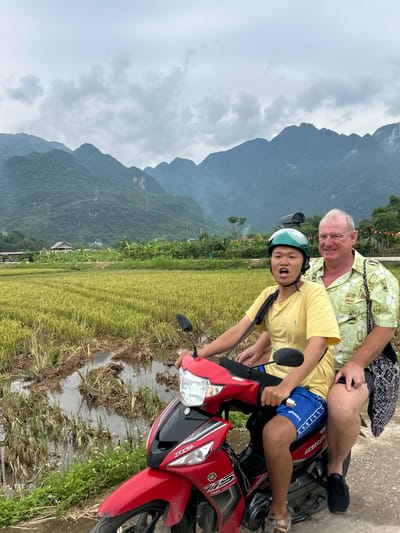 In Mai Chau, Vietnam, two individuals are depicted riding a red motorcycle on a dirt path beside a rice paddy. The person in front, wearing a green helmet, a yellow t-shirt, and blue shorts, appears to be a young male. He is leaning forward with his mouth open. The person behind him, a mature male with light skin and short hair, is wearing a light green patterned shirt and black shorts. He has his arm around the younger individual. The background features lush green rice fields, a cluster of banana trees, and mist-covered mountains under a cloudy sky. A small thatched-roof structure is visible in the middle ground. The overall scene suggests a rural and natural environment.