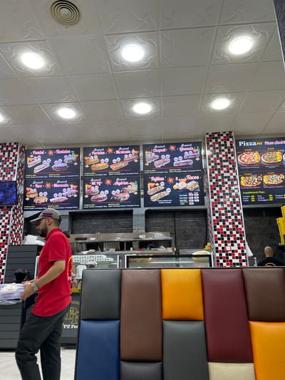 The image captures the interior of a fast-food establishment named "Pif Food" located in Baraki, Algeria. The scene is dominated by a bright, fluorescent-lit ceiling with square tiles. On the walls, there are large menu boards displaying various food items like paninis, sandwiches, fajitas, tacos, and pizzas, each with prices listed in Algerian Dinars. The menus feature images of the food and descriptive text in French and Arabic.
In the foreground, a man wearing a red polo shirt and a patterned baseball cap is seen from the side, carrying a tray with food items. He appears to be a staff member, as suggested by his attire and action of transporting food. Behind him, a counter area is visible with food preparation equipment and displays. To the right, a row of colorful padded seating is arranged, consisting of brown, dark gray, blue, burgundy, orange, and beige panels. In the background, other customers and staff are partially visible. The overall environment suggests a busy, casual dining setting. The decor includes checkered patterns in red, black, and white on some of the vertical surfaces.
