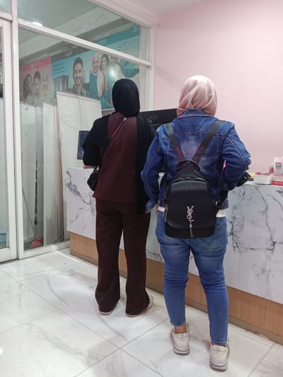 Two women are standing back-to-back in what appears to be a beauty salon or cosmetic store. The woman on the left is wearing a black hijab and a maroon top and pants. She has a small black purse slung over her shoulder. The woman on the right is wearing a light pink hijab and a denim jacket over a white t-shirt, paired with blue jeans. She is carrying a black Yves Saint Laurent backpack.
The background shows a counter with a marble-like pattern and some products on display. Behind the counter, there's a computer monitor. A large poster is visible through a glass partition on the left, featuring images of people and text in what appears to be Indonesian. One poster segment has text that reads "#BEAUTY MOVES YOU". The lighting suggests it's daytime, and the indoor setting implies no specific weather conditions are relevant. The overall mood is neutral, with the women appearing to be engaged in some transaction or browsing. The floor is tiled with a similar marble pattern. The location context is Kaliwungu, Indonesia.