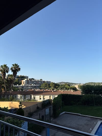 The image displays a panoramic view of a coastal town, likely Sant Feliu de Guíxols, Spain. The scene is bathed in the clear, bright light of a sunny day. The sky is a vibrant, cloudless blue, occupying a significant portion of the left side of the frame.
In the mid-ground and stretching into the background, a densely built urban landscape is visible. Buildings of various heights and architectural styles, many with terracotta tiled roofs, are nestled on a gentle hillside. Some buildings appear to be apartment complexes with numerous balconies, while others are smaller, more traditional structures. Lush green trees and vegetation are interspersed throughout the town, softening the urban environment.
In the foreground on the right, a swimming pool with blue water is partially visible, bordered by a tiled patio and a low wall. Adjacent to the patio is a patch of neatly mown green grass, and further back, a dense hedge lines the property. A metal railing runs along the right edge of the image, suggesting the viewpoint is from a balcony or elevated terrace.
On the left side of the foreground, a dark, angled structure, possibly a roof or awning, partially obscures the view of the sky. In the middle ground, several palm trees with their distinctive fronds are prominent, indicating a warm, Mediterranean climate. The overall impression is one of a serene and pleasant day in a well-established residential area overlooking a town and possibly the sea, though the sea itself is not directly visible. The time of day appears to be late afternoon or early evening, given the quality of the light. No people or specific activities are depicted, and there is no visible text.