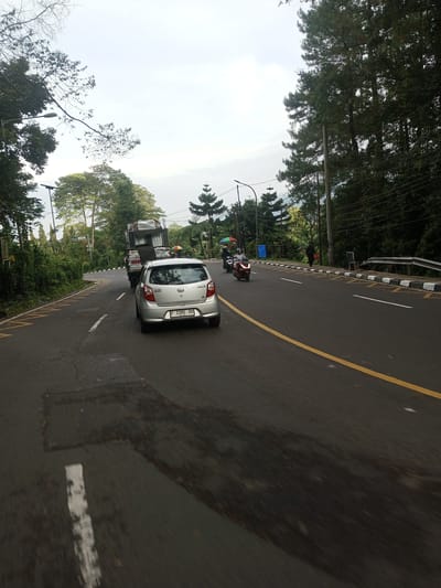 The image shows a road in a hilly, wooded area. A silver car is in the left lane, followed by a truck. Two motorcycles are in the right lane. A person is standing on the sidewalk. Trees line both sides of the road. The sky is overcast. The license plate of the car is partially visible as F 1180 XV. The road has yellow and white lane markings. The image's location is NOT in Taunggyi, Myanmar.