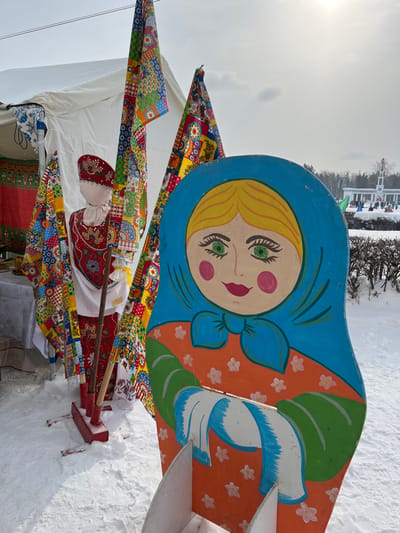 The image captures an outdoor scene during what appears to be a winter festival or cultural event. The ground is covered in a thick layer of snow, indicating cold weather. The sky is bright but overcast, suggesting a cloudy day.
In the foreground, a large, brightly painted wooden cutout of a Matryoshka doll (Russian nesting doll) stands prominently. The doll's headscarf is blue, framing a face with blonde hair, green eyes, pink rosy cheeks, and red lips. Its body is painted orange with delicate white and green floral patterns, and there are distinct white and blue elements at its base, resembling a sash or apron.
To the left and slightly in front of the large Matryoshka, a smaller, traditional-looking effigy or doll is visible, propped up on a wooden pole. This figure is dressed in red and white clothing with intricate patterns, including a red headpiece.
Numerous colorful flags or banners are displayed around these figures. These banners feature a vibrant patchwork design with various geometric and floral patterns in blues, reds, yellows, and greens, contributing to a festive atmosphere. They are attached to poles, some of which are held by the smaller effigy.
In the background, beyond the immediate display, there is a white tent or canopy. Further back, a line of trees, likely snow-covered, stretches across the landscape, and a distinct white building with classical-style pillars or columns can be seen, suggesting a park or estate setting.
The overall impression is one of a cheerful and colorful display, likely part of a public celebration or fair, possibly with Russian cultural themes given the Matryoshka doll. No discernible text is visible in the image.