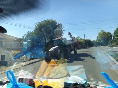 The image provides a view from inside a vehicle, looking out through a windshield covered in smudges and reflections. In the middle ground, a large agricultural tractor with a red attachment is visible on a paved road. The tractor is silver and grey, with large rear wheels. The road curves to the right, with a curb and vegetation on the right side. The sky is clear blue, with several power lines stretching across the frame. On the left side of the road, a building with a tiled roof is visible. The foreground of the image is cluttered with various items, including blue plastic bags, rolls of toilet paper, and yellow cylindrical objects, all appearing to be inside the vehicle. The scene is set in Krumovo, Bulgaria, indicated by the presence of a stop sign on the right side of the road. The overall impression is a sunny day, likely in a rural or semi-rural area.