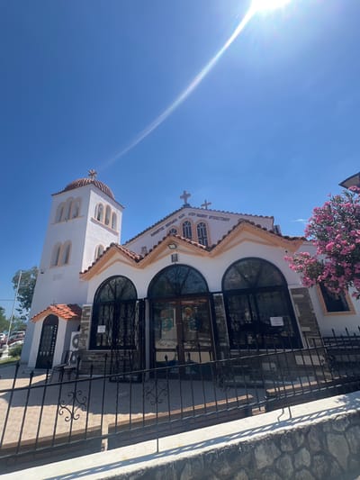 This image captures a brightly lit exterior of a white church with a red-tiled roof under a clear blue sky. The church features a prominent bell tower with arched windows and a domed roof topped with a cross. The main building has a series of arched windows and doors, some with ornate black metalwork. A pink flowering bush is visible on the right side of the church. The foreground shows a black metal fence and a stone wall. Inscriptions in Greek are visible on the church facade. Based on the text, the church is identifiable as the Panagitsa Monastery of Agios Ioannis Chrysostomos (Holy Monastery of Saint John Chrysostom), located in the municipality of Pallini, in the Attica region of Greece. There are no people prominently featured in the image.