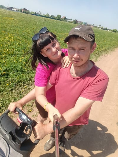 A couple is taking a selfie on a dirt path near a field in Foki, Russia. The man, wearing a red t-shirt and a Puma baseball cap, is holding the selfie stick. The woman, in a pink t-shirt and with sunglasses on her head, has her hand on his shoulder. They are near a stroller. In the background is a field with yellow flowers and houses.