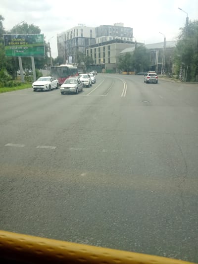 The image shows a street scene in Taraz city administration, Kazakhstan. Several cars are visible on the road, including a white SUV, several white sedans, and a red and white bus. The cars are driving in the same direction. A billboard with text in Kazakh and Russian is visible on the left side of the image, along with trees and buildings in the background. The sky is overcast. The image appears to have been taken from inside a vehicle.