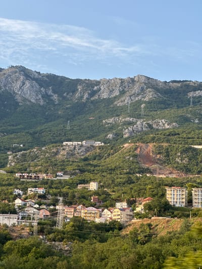 The image depicts a hillside residential area in Markovići, Montenegro, under a clear blue sky with wispy clouds. The landscape is characterized by a steep mountain covered in dense green foliage, with scattered rocky outcrops. Several buildings are visible on the slopes, ranging from multi-story apartment complexes to smaller houses. Some of these structures appear to be under construction or renovation, indicated by exposed materials and scaffolding. Electricity pylons are also present, running up the mountainside, suggesting the presence of power infrastructure. The foreground is dominated by lush green trees and vegetation. The scene conveys a sense of a developing community nestled within a natural, mountainous environment.