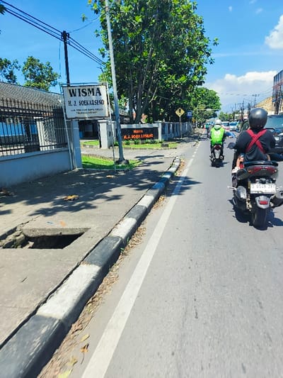 The image shows a street scene in Kecamatan Margahayu, Indonesia, taken from a moving vehicle. The street is paved with asphalt and has a white dividing line. There is a sidewalk on the left side of the street, lined with a concrete curb and a row of trees. A sign for "WISMA H.J. SOEKARSO" is visible at the beginning of the sidewalk, indicating a building or complex nearby. The image shows a motorcycle in the foreground, while several other motorcycles and a car are visible in the distance, moving in the same direction. The sky is clear and blue with white clouds. The scene is a typical street environment, with a mix of residential and commercial properties. The time of day is likely early afternoon.  The photo appears to have been taken from the back of a motorcycle or scooter, as seen by the blur of the street and the visible rider. The image captures a moment of everyday life in Kecamatan Margahayu, Indonesia. The coordinates (latitude: -6.981, longitude: 107.562) provide a precise location for this scene.
