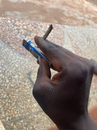A dark-skinned hand holds a lit cigarette or joint and a blue lighter. The hand's fingers are curled around the lighter, positioning it to ignite the cigarette. The cigarette is short and appears partially burned. The background shows a patterned tile or stone surface. The focus is sharp on the hand and its contents, with a slight blur to the background.