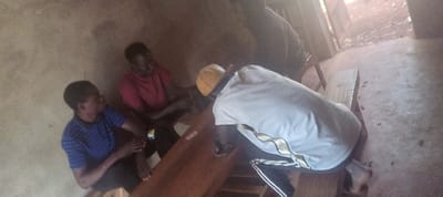 The scene, located in Getenga ward, Kenya, depicts an indoor environment with rough, light-colored walls and a grey concrete floor. Four adult males are present. A large, smooth, dark wooden beam or log lies horizontally across the midground. One male, wearing a light-colored t-shirt with dark stripes and dark track pants, and a light-yellow cap or hard hat, is bent over the wooden beam, hands engaged with it. To his left, a male in a red t-shirt is seated or kneeling, also engaging with the beam. Further left, a male in a blue striped polo shirt is seated or kneeling, observing or assisting. A fourth male in a dark shirt is partially visible in the upper center, near the beam. Natural light enters from an opening on the right, illuminating the concrete floor where small dark debris is present. A narrow, light-colored wooden plank or bench is visible on the right foreground.