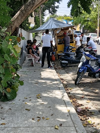 The image depicts a street scene in Puerto Plata, Dominican Republic, with several people gathered around a food stand. The day appears to be sunny, casting shadows on the concrete sidewalk.
The main subjects are a group of people, some standing and some sitting in chairs. They are gathered near a yellow food cart, which is covered with a silver tarp. Several motorcycles are parked nearby, suggesting the area is a common gathering spot or thoroughfare.
The people are casually dressed, and some appear to be interacting with the food vendor or other individuals. One person is standing and actively engaged with the cart, possibly purchasing food. Others are seated, observing or waiting. The atmosphere seems relaxed, with people enjoying the outdoors.
Behind the food stand, a large tree with lush green foliage is visible, providing shade. In the background, there are glimpses of buildings and other vehicles, indicating an urban or semi-urban setting. The sidewalk is made of concrete and is partially covered with fallen leaves, adding to the natural elements of the scene.
There is no visible text in the image that can be easily deciphered. The time of day is likely afternoon, given the angle of the shadows. The overall impression is a snapshot of everyday life in a tropical environment, with a focus on a local food vendor and the community around it.