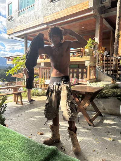 An outdoor scene in Pai, Thailand, depicts a young adult male posing shirtless in the foreground. He has dark, curly hair and is flexing his biceps, holding a black garment in his left hand. He wears black boxer briefs, tan/camouflage cargo shorts with a black belt, and tan combat-style boots. A necklace with a white, pointed pendant hangs from his neck. A faint tattoo is visible on his left torso.
To the left, another individual is partially visible from the lower back down, wearing black shorts and black flip-flops. In the background, a multi-story building constructed with grey concrete-like walls on the upper level and wooden structures below serves as the primary backdrop. This building features multiple wooden balconies, some with potted plants, and a distinctive metal spiral staircase with wooden treads on the right. Wooden doors with decorative panels are visible on the ground floor.
The ground is paved with grey interlocking pavers. To the left, wooden picnic-style benches and tables are arranged. To the right of the main subject stands a small, foldable wooden table. Lush green foliage and distant mountains are visible under a partly cloudy blue sky, suggesting an open landscape. An artificial green turf mat occupies the lower-left foreground. The scene is illuminated by bright natural light, casting shadows.
