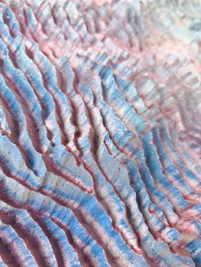 The image shows a close-up, abstract view of a textured surface with undulating ridges. The surface is colored with streaks of light blue and pink, interspersed with white and some darker speckles. The texture appears rough and granular, with the ridges casting subtle shadows, suggesting a light source from above and to the side.
The overall impression is of a natural, weathered surface, possibly sand dunes or a rock formation, with the colors suggesting a painted or mineralized appearance. The angle of the shot and the depth of field create a sense of scale and texture. There are no discernible subjects like people or objects, nor any identifiable scene or setting beyond the immediate textured surface. No activity or event is taking place, and no time of day, weather, or location cues are evident. There is no visible text in the image. The abstract nature of the image focuses on color and texture.