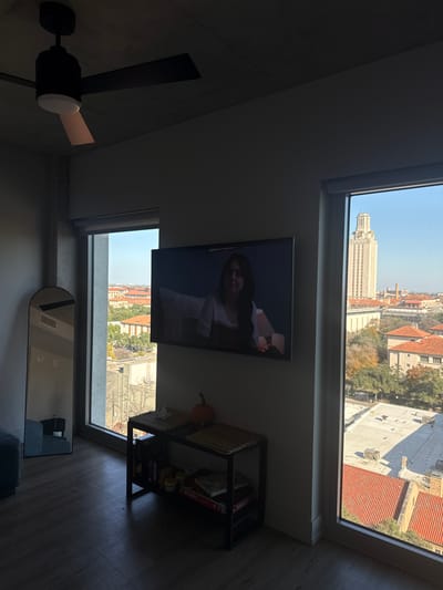 The image, appearing rotated 90 degrees counter-clockwise, depicts an indoor room with a prominent view of a city through large windows. When correctly oriented, the room features light-colored walls and a light wood-toned floor.

In the foreground, a black ceiling fan with a white central light fixture hangs from the ceiling. On the far wall, a flat-screen television is mounted, displaying a clear image of a woman with long dark hair, wearing a light top, looking towards the viewer. To the right of the television, positioned against the wall, is a dark-framed shelf unit with two levels. The top shelf holds woven baskets, one of which contains a small, orange decorative pumpkin. The bottom shelf is filled with board games, with the word "BLIZZARD" clearly visible on one box and possibly "PICTIONARY" on another. Below the right window, a large, dark-framed mirror with a curved top is leaning against the wall, reflecting parts of the room.

Through the expansive windows, a bright, clear daytime sky is visible. The outdoor scene reveals a city skyline, dominated by a tall, light-colored architectural building featuring a distinct tiered or domed tower, strongly resembling the Main Building and Tower of the University of Texas at Austin. Surrounding this landmark are numerous other city buildings, some with distinctive red-tiled roofs and others with flat, grey rooftops. Scattered trees with hints of autumnal foliage are also visible among the buildings. The clear sky and bright outdoor light suggest a sunny day. The pumpkin decoration suggests the image was taken around autumn or Halloween.