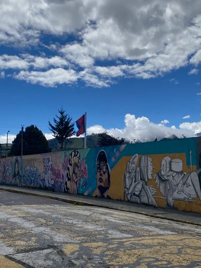 The image shows a vibrant mural on a wall next to a patterned sidewalk, under a bright blue sky with scattered clouds. A tree and a flag of Quito, Ecuador, stand in front of a distant hill. The mural has several distinct sections, including stylized portraits and abstract designs. The location is likely Quito, Ecuador, as indicated by the flag. The scene appears to be a street view in a neighborhood known for street art. There are no people visible in the image.