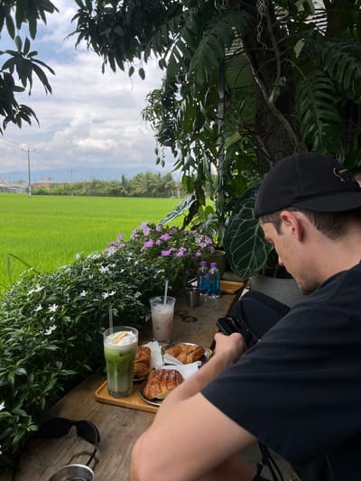 A man in a black shirt and cap is seated at a wooden table with a view overlooking a rice field. The table is set with croissants on a tray, two drinks, small bottles of Okado mineral water, and sunglasses. The scene is framed by foliage with white and purple flowers. The location is implied to be in Diên Khánh, Vietnam, based on the rice paddy landscape. The man is looking down at his phone, which he is holding in his hand.