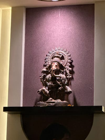 A bronze-colored, ornate depiction of Ganesha is centered on a textured, plum-colored surface. Ganesha is intricately detailed with multiple figures and a circular, sun-like halo. A dark, rounded shape with a black rectangular frame partially obscures the right side of the image. The entire scene is lit from the bottom, with a diffused, indirect light casting shadows and highlighting the textures.