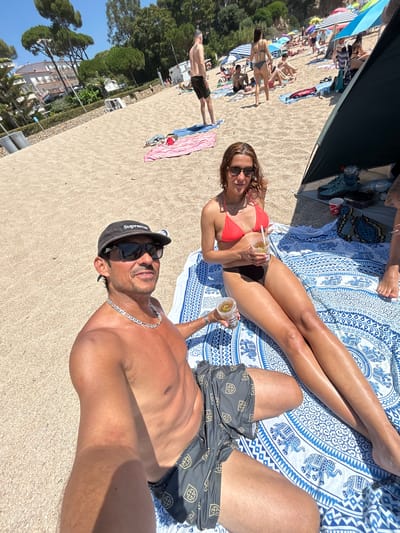 In Castell d'Aro, Platja d'Aro i s'Agaró, Spain, a couple is relaxing on a sandy beach. The man, wearing a black baseball cap and sunglasses, is in the foreground, facing the camera with his torso bare. He is wearing patterned swim trunks and a silver chain necklace. Next to him, a woman is seated on a blue and white patterned blanket, wearing a coral bikini top and black bottoms. She is holding a drink with a straw and looking towards the camera. Her legs are stretched out on the blanket. The background shows a bustling beach scene with other people, umbrellas, and tents. Lush green trees and buildings are visible behind the beach, suggesting a coastal town setting. The sky is clear and blue, indicating a sunny day.