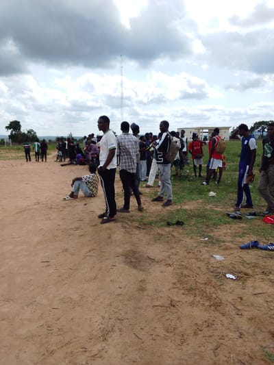 In Kachia, Nigeria, a group of young men and boys are gathered on a dirt field under a cloudy sky. The scene depicts an informal outdoor gathering, possibly a sports event or a casual meetup. The individuals are dressed in a mix of casual wear, including t-shirts, shorts, and pants. Some are standing, while others are seated or reclining on the ground. The ground is a mixture of dirt and sparse vegetation. In the background, there are trees, a building with a white facade, and a tall antenna tower. The overall atmosphere appears relaxed, with a sense of community.