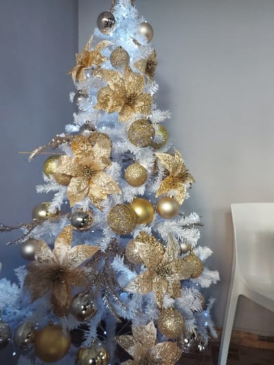 The image captures a close-up, horizontal view of a beautifully decorated white Christmas tree. The tree's white faux pine branches are heavily adorned with an elegant array of gold and silver ornaments. Prominently featured are numerous spherical golden baubles, varying in texture from smooth and matte to glitter-covered and intricately patterned. These are interspersed with metallic silver or champagne-colored spheres. Large, glistening golden flowers, resembling poinsettias, are strategically placed among the branches, contributing significant sparkle. White LED string lights are woven throughout the tree, casting a soft, bright glow that accentuates the festive decorations.
The tree is situated indoors, set against a plain light gray wall, which provides a neutral backdrop for the vibrant ornaments. In the upper right corner, a glimpse of a white, modern-looking ceiling fixture or appliance is visible. The scene depicts a holiday decoration, creating a festive and elegant atmosphere, likely during the Christmas season. No visible text is present in the image. This festive setup is located in Tiranë, Albania.